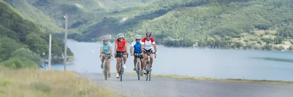 Quatre cyclotouristes, deux hommes et deux femmes, roulent à vélo au bord d’un lac sur une route légèrement en montée, sourires aux lèvres.